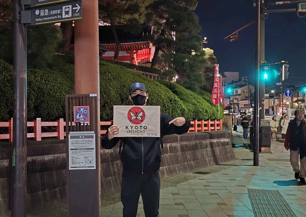 A Kyoto Insight guide holding a sign at the illuminated entrance of Yasaka Jinja Shrine, welcoming visitors to Kyoto free walking tours heading to the gion geisha district through higashiyama